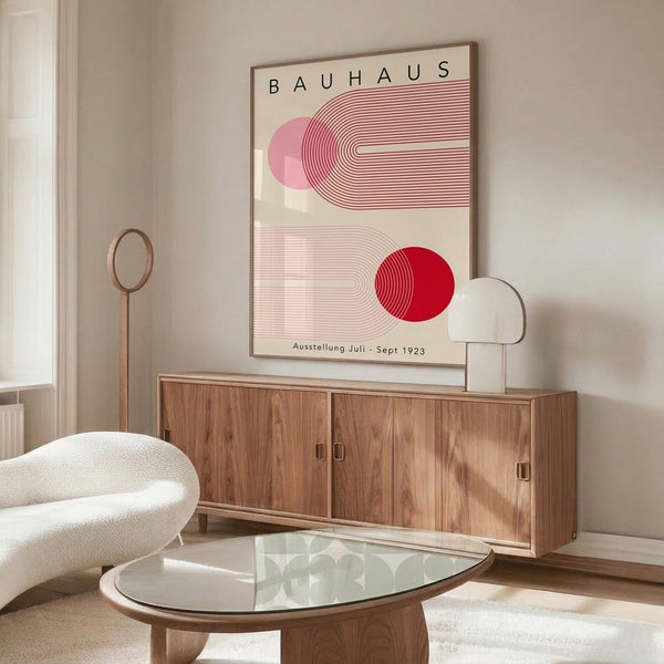 May include: Bauhaus exhibition poster from 1923 with geometric red and pink semicircular designs and linear patterns, displayed above a wooden mid-century modern credenza in a minimalist living room with cream walls, white sculptural chair, glass coffee table, and modern lamp