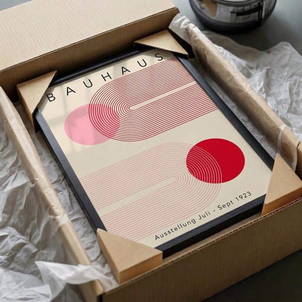 May include: Framed Bauhaus exhibition poster with geometric design featuring pink and red circles with curved line patterns, displayed in black frame inside cardboard shipping box with protective wrapping materials on white surface