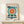 May include: Framed Bauhaus-style poster with geometric design featuring a large teal circle containing radiating red and white sunburst pattern with yellow center, angular black architectural elements below, and "BAUHAUS" text at top. Natural wood frame displayed on light wooden surface next to small white wrapped gift with green sprig, against cream wall.