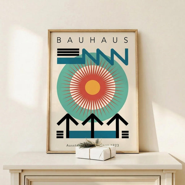 May include: Framed Bauhaus-style poster with geometric design featuring a large teal circle containing radiating red and white sunburst pattern with yellow center, angular black architectural elements below, and "BAUHAUS" text at top. Natural wood frame displayed on light wooden surface next to small white wrapped gift with green sprig, against cream wall.
