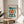 May include: Framed Bauhaus exhibition poster with geometric design featuring large teal circle, radiating red and yellow sunburst center, black zigzag arrows, and horizontal lines, displayed on round wooden stool next to beige upholstered chair with plant shadow on white wall