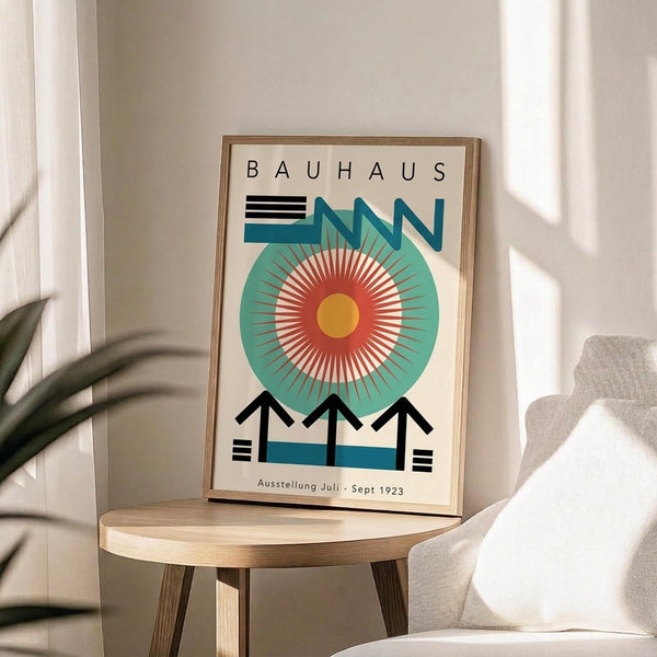 May include: Framed Bauhaus exhibition poster with geometric design featuring large teal circle, radiating red and yellow sunburst center, black zigzag arrows, and horizontal lines, displayed on round wooden stool next to beige upholstered chair with plant shadow on white wall