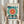 May include: Person in cream sweater holding framed Bauhaus exhibition poster featuring geometric design with large teal circle, radiating orange and red center, black architectural elements, and "Ausstellung Juli - Sept 1923" text. Light wood frame against white curtained background with dried pampas grass visible on left side.