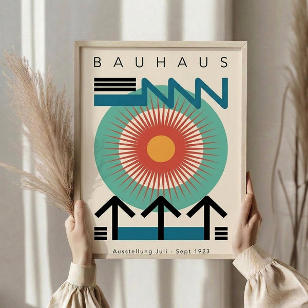 May include: Person in cream sweater holding framed Bauhaus exhibition poster featuring geometric design with large teal circle, radiating orange and red center, black architectural elements, and "Ausstellung Juli - Sept 1923" text. Light wood frame against white curtained background with dried pampas grass visible on left side.