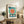 May include: Framed Bauhaus exhibition poster with geometric design featuring large teal circle, radiating orange and red sunburst pattern, black angular shapes, and "BAUHAUS" text at top. Displayed on rustic wooden sideboard alongside fern plant, ceramic vase, and books in neutral-toned living room setting.