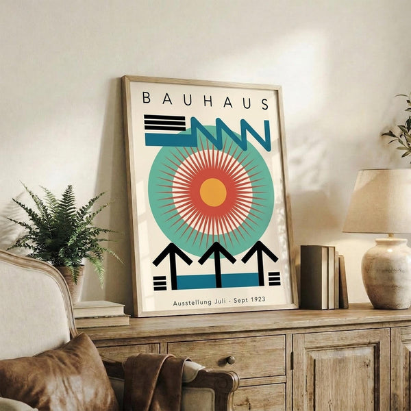 May include: Framed Bauhaus exhibition poster with geometric design featuring large teal circle, radiating orange and red sunburst pattern, black angular shapes, and "BAUHAUS" text at top. Displayed on rustic wooden sideboard alongside fern plant, ceramic vase, and books in neutral-toned living room setting.