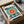 May include: Hands unboxing a framed Bauhaus exhibition poster from 1923 featuring a stylized sunburst design with concentric circles in teal, coral red, and yellow. The geometric artwork displays "BAUHAUS" and "Ausstellung Juli - Sept 1923" text with bold black triangular elements. Black frame shown partially unwrapped in cardboard shipping box with white tissue paper on marble surface.