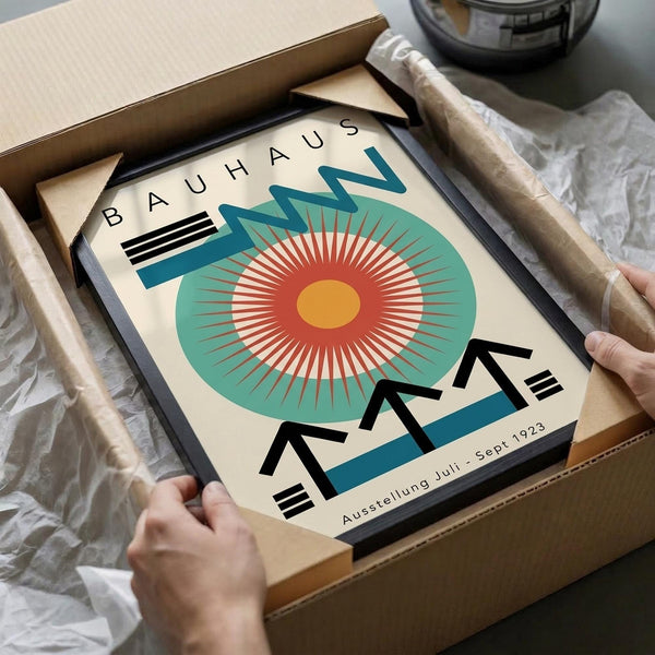 May include: Hands unboxing a framed Bauhaus exhibition poster from 1923 featuring a stylized sunburst design with concentric circles in teal, coral red, and yellow. The geometric artwork displays "BAUHAUS" and "Ausstellung Juli - Sept 1923" text with bold black triangular elements. Black frame shown partially unwrapped in cardboard shipping box with white tissue paper on marble surface.