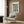 May include: Modern living room scene featuring a framed Bauhaus exhibition poster on wood-paneled wall. The colorful geometric poster displays a radiating sunburst pattern in teal, coral, and yellow with angular architectural elements and German text. Below sits a cream upholstered armchair with patterned throw pillow, brass floor lamp, and dark round side table with open book. Natural light filters through curtained window.