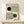 May include: Framed Bauhaus-style poster with geometric design featuring black and green circles intersected by flowing concentric lines on cream background, displayed on light wood shelf next to small wrapped gift, leaning against white wall in minimalist room setting.