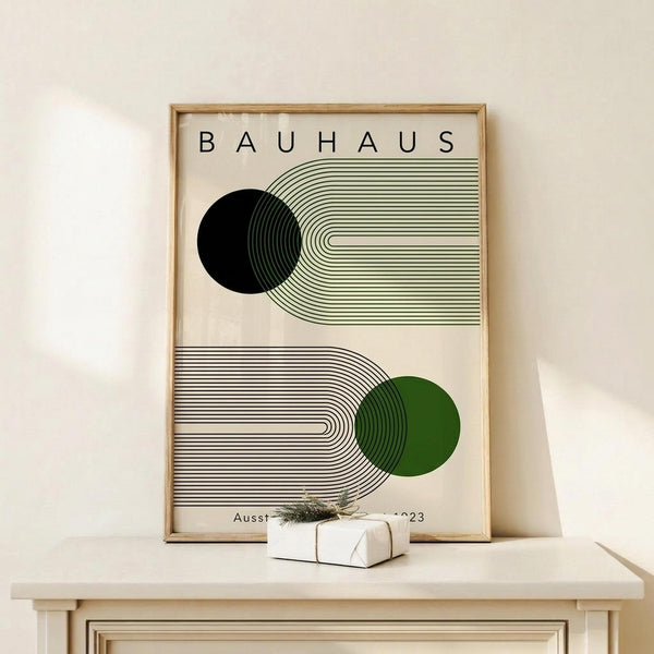 May include: Framed Bauhaus-style poster with geometric design featuring black and green circles intersected by flowing concentric lines on cream background, displayed on light wood shelf next to small wrapped gift, leaning against white wall in minimalist room setting.