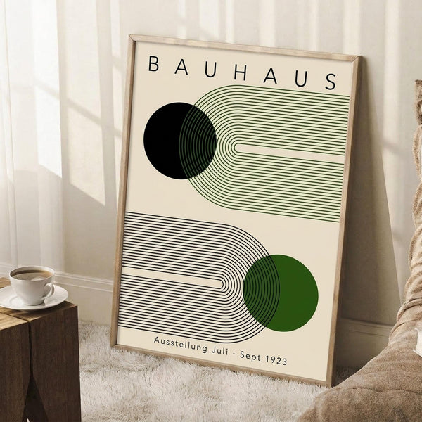 May include: Framed Bauhaus-style poster with geometric design featuring flowing curved lines in green and black, with black and green circles. The minimalist artwork displays "BAUHAUS" at top and "Ausstellung Juli - Sept 1923" at bottom on cream background. Light wood frame leans against white wall corner next to wooden stool with white coffee cup and saucer, positioned on textured cream rug in bright natural lighting.