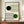 May include: A framed Bauhaus-style poster with geometric black and green circles intersecting curved parallel lines on cream background, displayed on a wooden sideboard next to a white ceramic vase and stacked books in a room with white paneled walls.