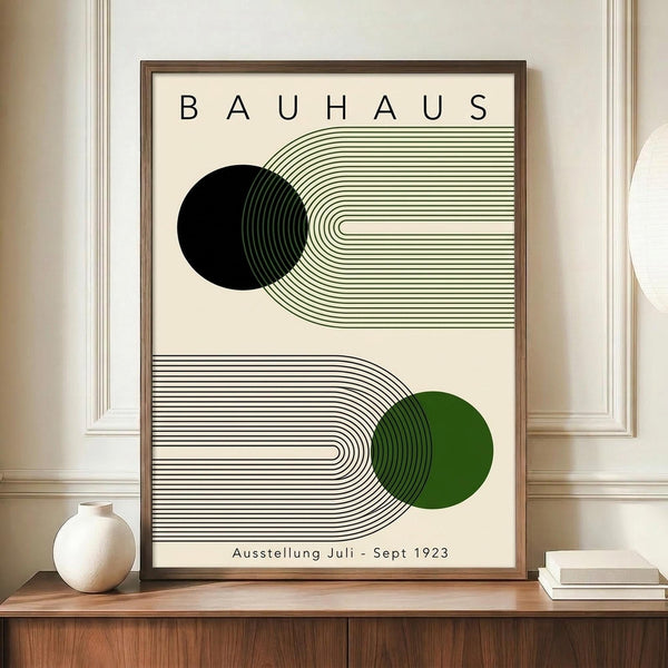 May include: A framed Bauhaus-style poster with geometric black and green circles intersecting curved parallel lines on cream background, displayed on a wooden sideboard next to a white ceramic vase and stacked books in a room with white paneled walls.
