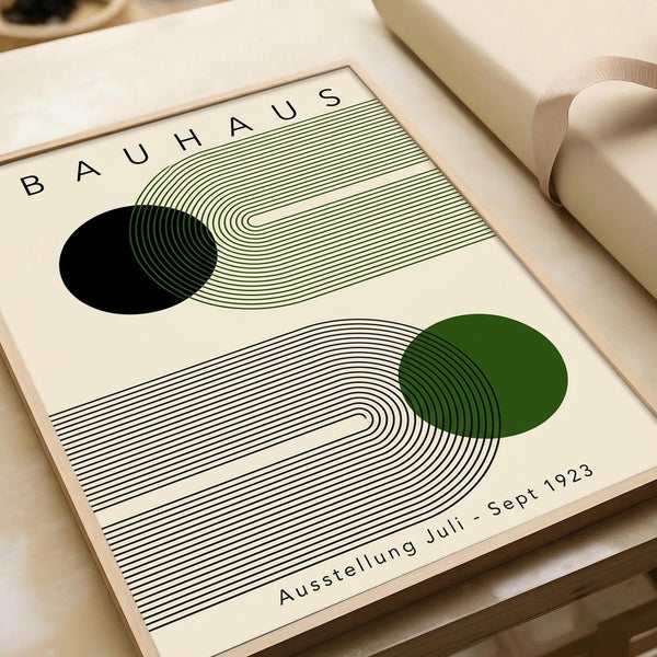 May include: Cream-colored poster print with abstract geometric design featuring flowing concentric lines in black and green, resembling topographical contours or sound waves. Two circular elements - one black, one forest green - intersect with the linear patterns. Text reads "BAUHAUS" at top and "Ausstellung Juli - Sept 1923" at bottom. Print appears matted and sits alongside neutral beige ribbon on light wooden surface.