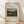May include: Framed art print featuring Henri Rousseau's 1895 pastoral landscape painting with four young girls in a green countryside scene, displayed in light wooden frame against white textured wall on wooden floor with natural window light creating soft shadows