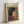 May include: Framed poster featuring a woman with dark hair in profile view wearing a red garment with decorative swirls, surrounded by colorful flowers on yellow background. The artwork displays Matisse-inspired cut-out style with bold colors including red, blue, and green florals. Natural wood frame sits against white wall, showing the cream-colored poster with "HENRI MATISSE" text at top and French subtitle.