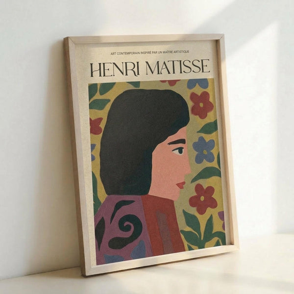 May include: Framed poster featuring a woman with dark hair in profile view wearing a red garment with decorative swirls, surrounded by colorful flowers on yellow background. The artwork displays Matisse-inspired cut-out style with bold colors including red, blue, and green florals. Natural wood frame sits against white wall, showing the cream-colored poster with "HENRI MATISSE" text at top and French subtitle.