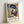 May include: Framed art print leaning against white wall showing colorful Fauve-style painting of woman in blue hat and floral dress beneath palm fronds, with Henri Matisse attribution text below artwork in natural wood frame