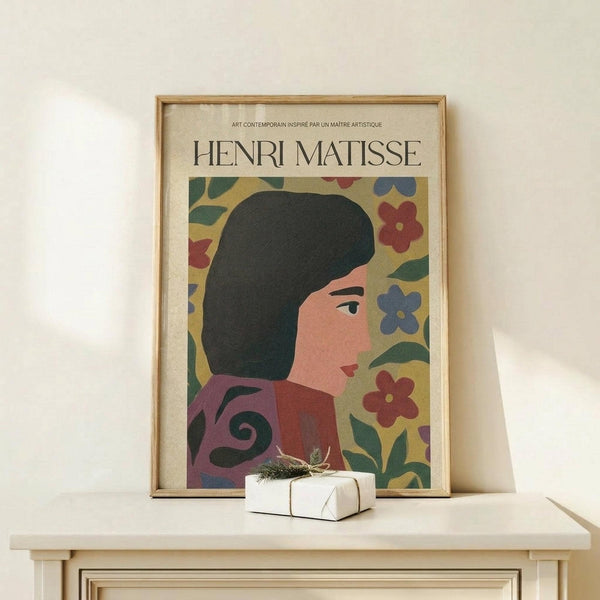 May include: Framed poster featuring a stylized woman with dark hair in profile view, surrounded by colorful flowers in red, blue, and green on yellow background. Contemporary art style with bold, flat colors and organic shapes. Light wood frame displayed on pale wooden surface with small wrapped gift box beside it against cream wall.