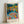 May include: Framed art print featuring a vibrant Fauve-style painting with bold brushstrokes in blues, oranges, yellows, and greens. The scene depicts figures gathered by a reflective body of water surrounded by colorful foliage. Text reads "HENRI MATISSE PARIS" at bottom. Light wooden frame sits propped against white wall on white surface.