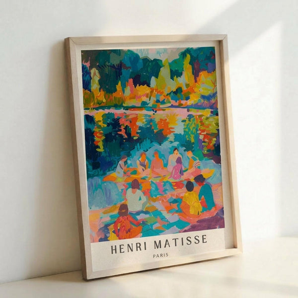 May include: Framed art print featuring a vibrant Fauve-style painting with bold brushstrokes in blues, oranges, yellows, and greens. The scene depicts figures gathered by a reflective body of water surrounded by colorful foliage. Text reads "HENRI MATISSE PARIS" at bottom. Light wooden frame sits propped against white wall on white surface.