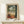 May include: Framed art print in natural wood frame showing tropical terrace scene with figure in orange dress on white balustrade, palm trees, small table with oranges, and Mediterranean landscape beyond archway. White wrapped gift with twine sits on light wood console table against cream wall.