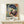 May include: Framed Henri Matisse exhibition poster featuring a woman in a wide blue hat and colorful floral dress holding a parasol, displayed on a light wood shelf against a cream wall. The vibrant artwork shows Matisse's characteristic bold colors - blues, oranges, greens, and pinks. A small white gift box with dried grass sits beside the thin natural wood frame.