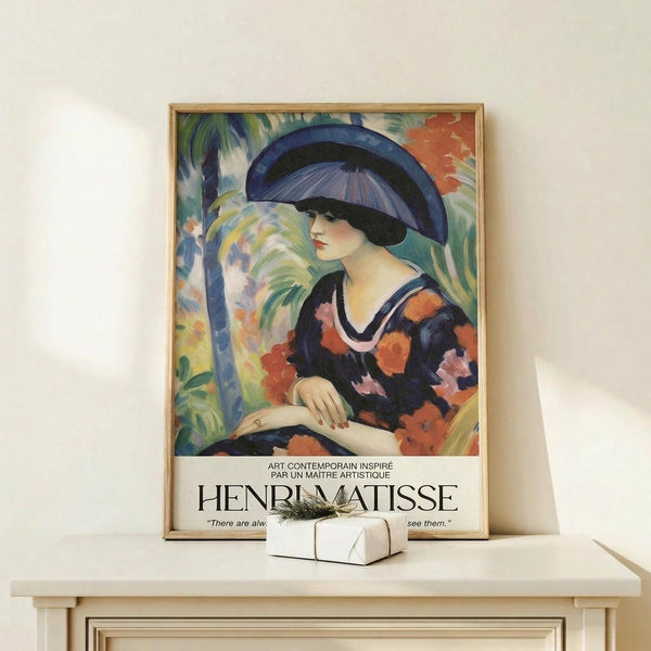 May include: Framed Henri Matisse exhibition poster featuring a woman in a wide blue hat and colorful floral dress holding a parasol, displayed on a light wood shelf against a cream wall. The vibrant artwork shows Matisse's characteristic bold colors - blues, oranges, greens, and pinks. A small white gift box with dried grass sits beside the thin natural wood frame.