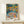 May include: Colorful Henri Matisse art print in thin wooden frame displayed on light wood console table against cream wall. Vibrant fauve-style painting shows figures by water with bold blues, oranges, yellows and greens. Small white wrapped gift with greenery sits beside the framed poster.