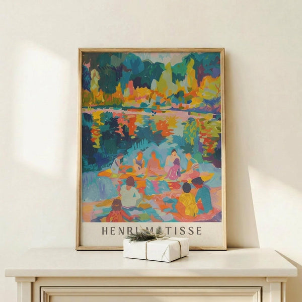 May include: Colorful Henri Matisse art print in thin wooden frame displayed on light wood console table against cream wall. Vibrant fauve-style painting shows figures by water with bold blues, oranges, yellows and greens. Small white wrapped gift with greenery sits beside the framed poster.