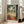 May include: Framed art print in light wood frame leaning against white wall next to beige sofa. Tropical scene shows woman in red dress on terrace overlooking palm trees and water, with fruit bowl on small table in foreground. Warm colors with green foliage framing archway view. Natural lighting from nearby windows creates soft shadows on wooden floor.