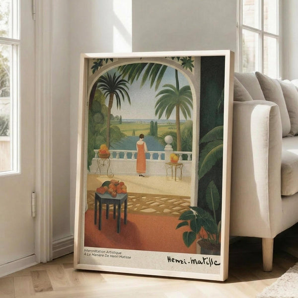 May include: Framed art print in light wood frame leaning against white wall next to beige sofa. Tropical scene shows woman in red dress on terrace overlooking palm trees and water, with fruit bowl on small table in foreground. Warm colors with green foliage framing archway view. Natural lighting from nearby windows creates soft shadows on wooden floor.