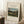 May include: A framed art print leaning against a white wall on a textured cream rug, showing Henri Rousseau's pastoral landscape painting with four young girls in a grassy meadow, trees, and industrial chimney smoke. A white coffee cup sits on a wooden block nearby, with soft natural lighting creating gentle shadows.