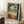 May include: Framed art print leaning against white wall on textured rug, showing tropical terrace scene with figure in red dress on balcony overlooking water, palm trees and lush greenery, ornate table with fruit bowl in foreground, coffee cup on wooden stool nearby, warm natural lighting creating soft shadows