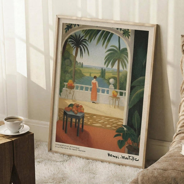 May include: Framed art print leaning against white wall on textured rug, showing tropical terrace scene with figure in red dress on balcony overlooking water, palm trees and lush greenery, ornate table with fruit bowl in foreground, coffee cup on wooden stool nearby, warm natural lighting creating soft shadows