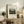 May include: Framed vintage art print of pastoral landscape with rolling green hills, trees, and small red figures in foreground, displayed on rustic wooden sideboard beside fern plant, books, ceramic vase, and beige armchair in warm-toned living room setting
