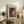 May include: Framed Henri Matisse-style poster displayed on rustic wooden sideboard in warm, neutral living room. Artwork features woman with dark bob haircut wearing red garment against colorful floral background with yellow, green, red and blue flowers. Natural wood frame, styled with fern plant, books, ceramic vase and cream upholstered chair with brown leather pillow.