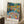 May include: Colorful Henri Matisse art print in thin wooden frame leaning against white wall corner. Vibrant Fauvist-style painting shows figures by water with bold blues, yellows, oranges, and greens. Artist name and Paris text at bottom. Room setting with white textured rug, wooden side table holding white coffee cup and saucer, beige knit throw blanket visible at edge.