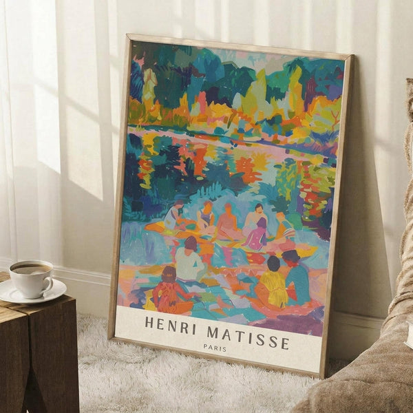 May include: Colorful Henri Matisse art print in thin wooden frame leaning against white wall corner. Vibrant Fauvist-style painting shows figures by water with bold blues, yellows, oranges, and greens. Artist name and Paris text at bottom. Room setting with white textured rug, wooden side table holding white coffee cup and saucer, beige knit throw blanket visible at edge.