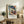 May include: Framed Henri Matisse poster print featuring a woman in a large blue hat and colorful floral dress against a vibrant tropical background with palm trees. The artwork is displayed on a rustic wooden sideboard alongside a potted fern, ceramic vase, and stacked books in a neutral-toned living room setting with cream upholstered furniture visible in the foreground.