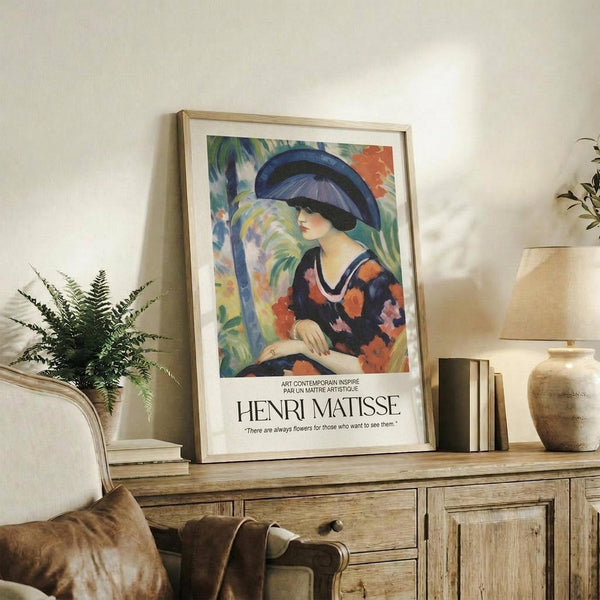 May include: Framed Henri Matisse poster print featuring a woman in a large blue hat and colorful floral dress against a vibrant tropical background with palm trees. The artwork is displayed on a rustic wooden sideboard alongside a potted fern, ceramic vase, and stacked books in a neutral-toned living room setting with cream upholstered furniture visible in the foreground.