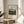 May include: Framed art print of Henri Rousseau's pastoral landscape painting featuring four young girls in a green countryside scene with rolling hills, trees, and buildings in the distance. The print has cream matting with black text and sits on a round wooden side table next to a beige upholstered chair. Soft natural lighting creates shadows from a nearby plant, giving the room a warm, minimalist aesthetic with neutral tones throughout.