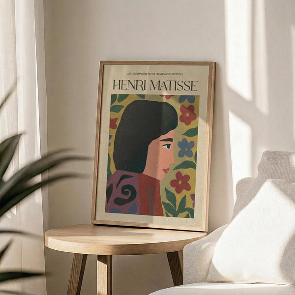 May include: Framed Henri Matisse-inspired poster featuring a woman with dark hair in profile against a vibrant floral background with red, blue, and yellow blooms. The colorful artwork sits on a light wood round side table next to a cream upholstered chair, with natural sunlight streaming across white walls and a potted plant visible in the corner of the minimalist room.