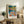 May include: Colorful framed art print displayed on rustic wooden sideboard in neutral living room setting. Vibrant artwork shows figures by water with autumn foliage in blues, oranges, yellows and greens. Natural wood frame with white matting. Scene includes fern plant, stacked books, ceramic vase, and cream upholstered furniture with brown leather pillow.