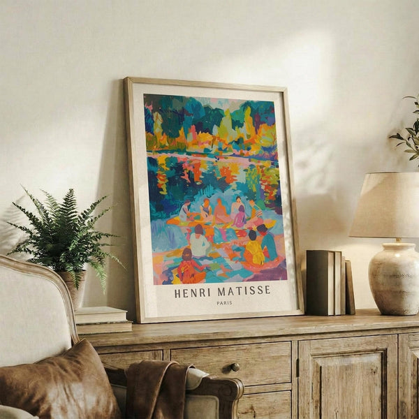 May include: Colorful framed art print displayed on rustic wooden sideboard in neutral living room setting. Vibrant artwork shows figures by water with autumn foliage in blues, oranges, yellows and greens. Natural wood frame with white matting. Scene includes fern plant, stacked books, ceramic vase, and cream upholstered furniture with brown leather pillow.