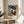 May include: Framed Henri Matisse art print displayed on round wooden side table next to beige upholstered chair. The colorful painting shows a woman in profile wearing a large blue hat and floral dress in vibrant oranges, pinks and blues against tropical foliage background. Natural sunlight casts shadows across the neutral-toned room with plant leaves visible in foreground.