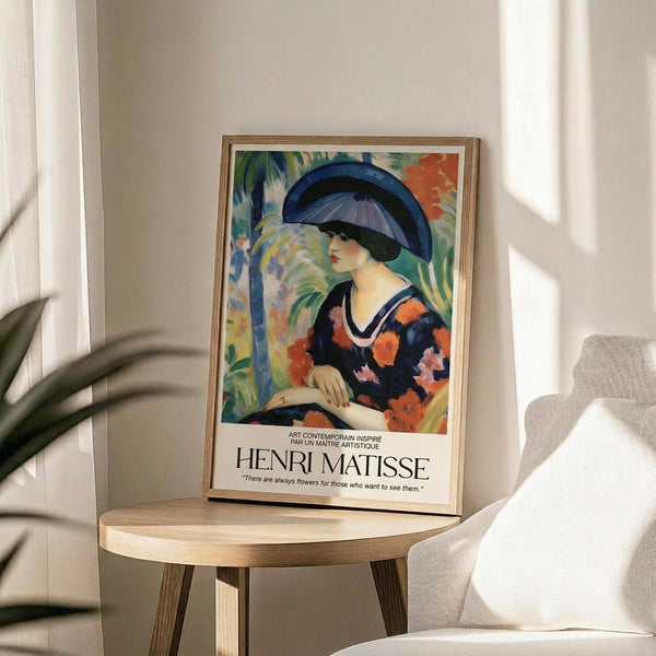May include: Framed Henri Matisse art print displayed on round wooden side table next to beige upholstered chair. The colorful painting shows a woman in profile wearing a large blue hat and floral dress in vibrant oranges, pinks and blues against tropical foliage background. Natural sunlight casts shadows across the neutral-toned room with plant leaves visible in foreground.