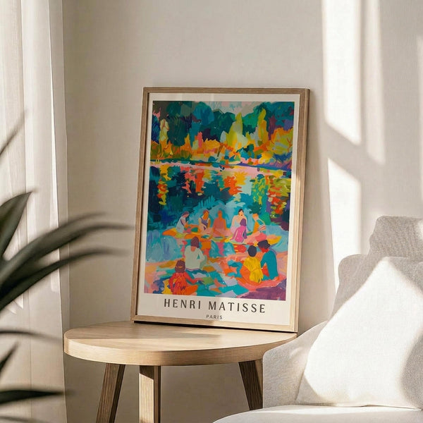 May include: Vibrant Henri Matisse poster print in thin gold frame, displayed on round wooden side table next to beige upholstered chair. Artwork features bold brushstrokes in blues, oranges, yellows, and pinks depicting figures by water with autumn foliage. Bright sunlight streams across white walls creating dramatic shadows from nearby plant leaves.