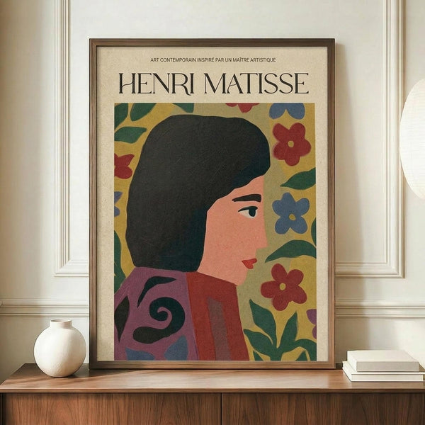 May include: Framed art print featuring a woman with dark hair and floral background in Matisse's cut-out style, displayed on wooden sideboard next to white ceramic vase and stacked books. The poster shows vibrant colors including yellow, red, blue, and green flowers with bold simplified forms. Brown wooden frame against cream colored wall with white trim molding.